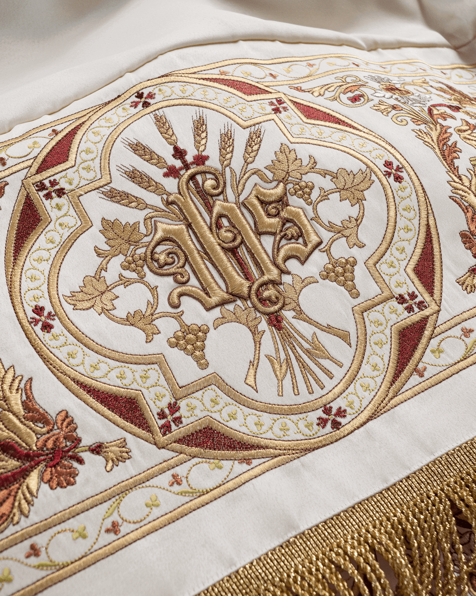 Haft monogramu IHS ze złotej nici, otoczony kłosami pszenicy i gronami winorośli, w czworolistnej ramie z czerwono-złotym ornamentem